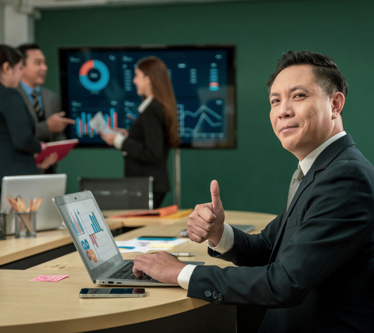 Picture of a team during a meeting, with someone sitting down, looking at the camera, and giving a thumbs up